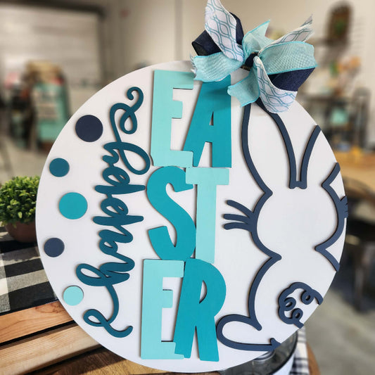 Decorative Easter sign with blue and white text and a bunny design, placed on a wooden surface.