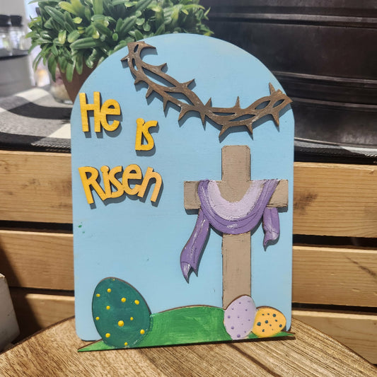 Decorative sign with 'He is Risen' text, cross, and Easter elements on a wooden surface.