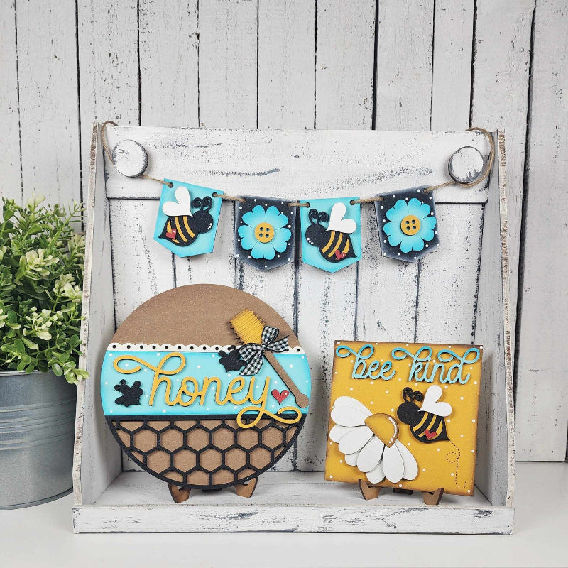 A decorative tiered tray set with a honeycomb design, bee and daisy decorations, and a 'Honey' sign, displayed against a rustic white background.