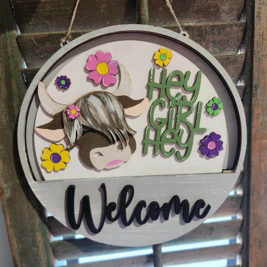 Decorative sign with highland cow, flowers, and 'Hey Girl Hey Welcome' text on a wooden background.