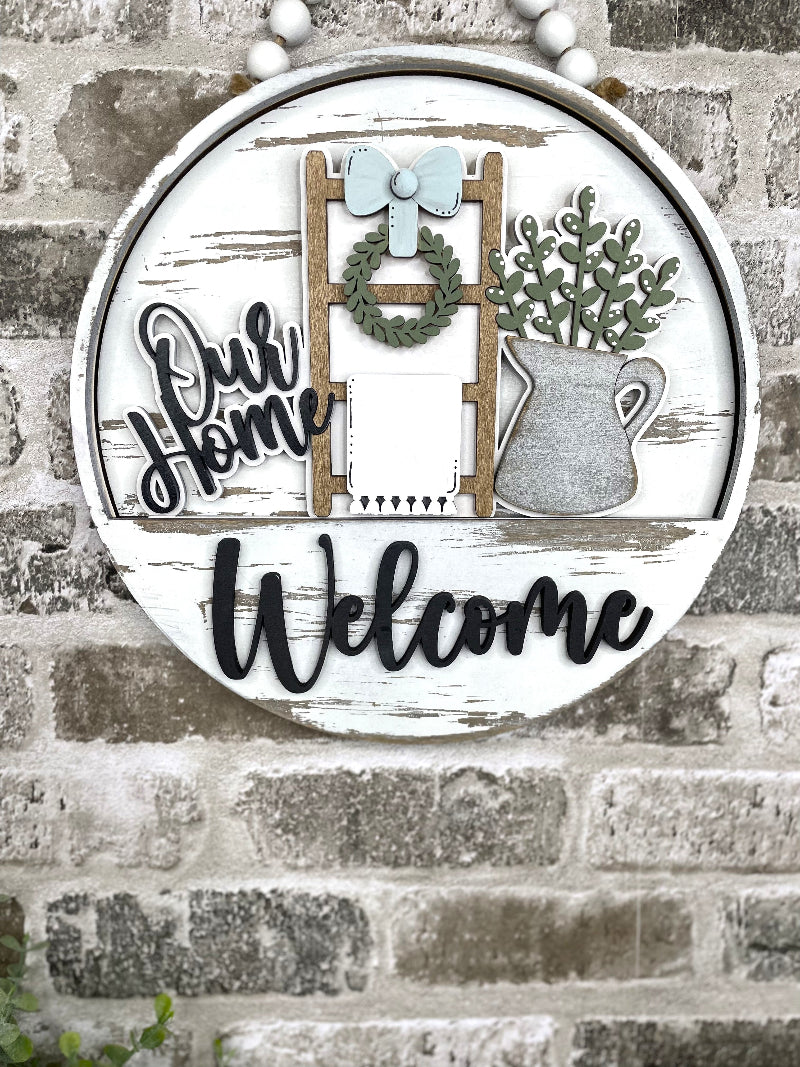 Our Home interchangeable wooden Welcome sign insert with farmhouse ladder, wreath, bow, hanging towel and metal pitcher filled with greenery.