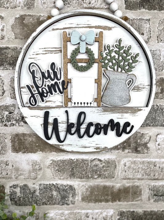 Our Home interchangeable wooden Welcome sign insert with farmhouse ladder, wreath, bow, hanging towel and metal pitcher filled with greenery.