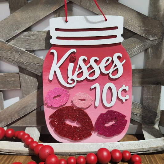 Decorative sign shaped like a pink jar with 'Kisses 10 cents' text, hanging against a wooden background.
