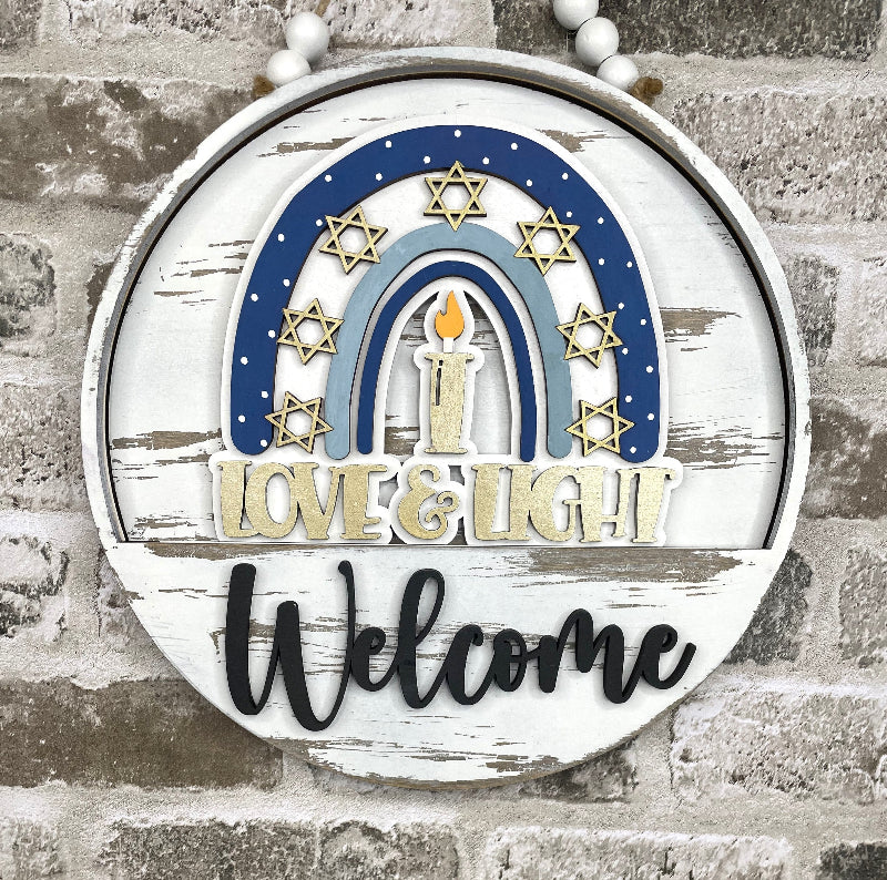 A round distressed white wooden sign with a blue and light blue rainbow, gold Star of David motifs, a central candle, and the words 'LOVE & LIGHT' and 'Welcome'. The sign hangs against a gray brick wall.