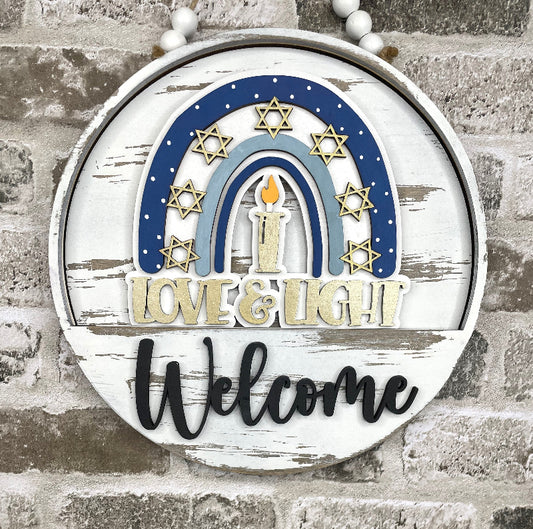 A round distressed white wooden sign with a blue and light blue rainbow, gold Star of David motifs, a central candle, and the words 'LOVE & LIGHT' and 'Welcome'. The sign hangs against a gray brick wall.