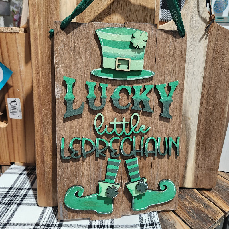 Wooden sign with green text 'Lucky little Leprechaun' and a green hat on a wooden surface.