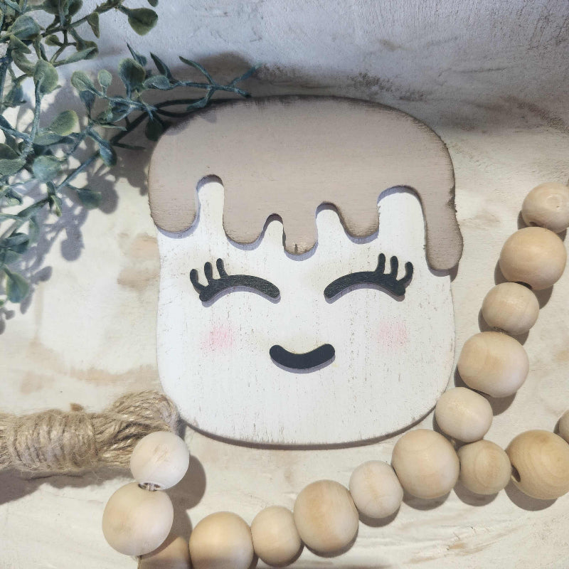 Decorative marshmellow with a face design and wooden beads on a textured surface.