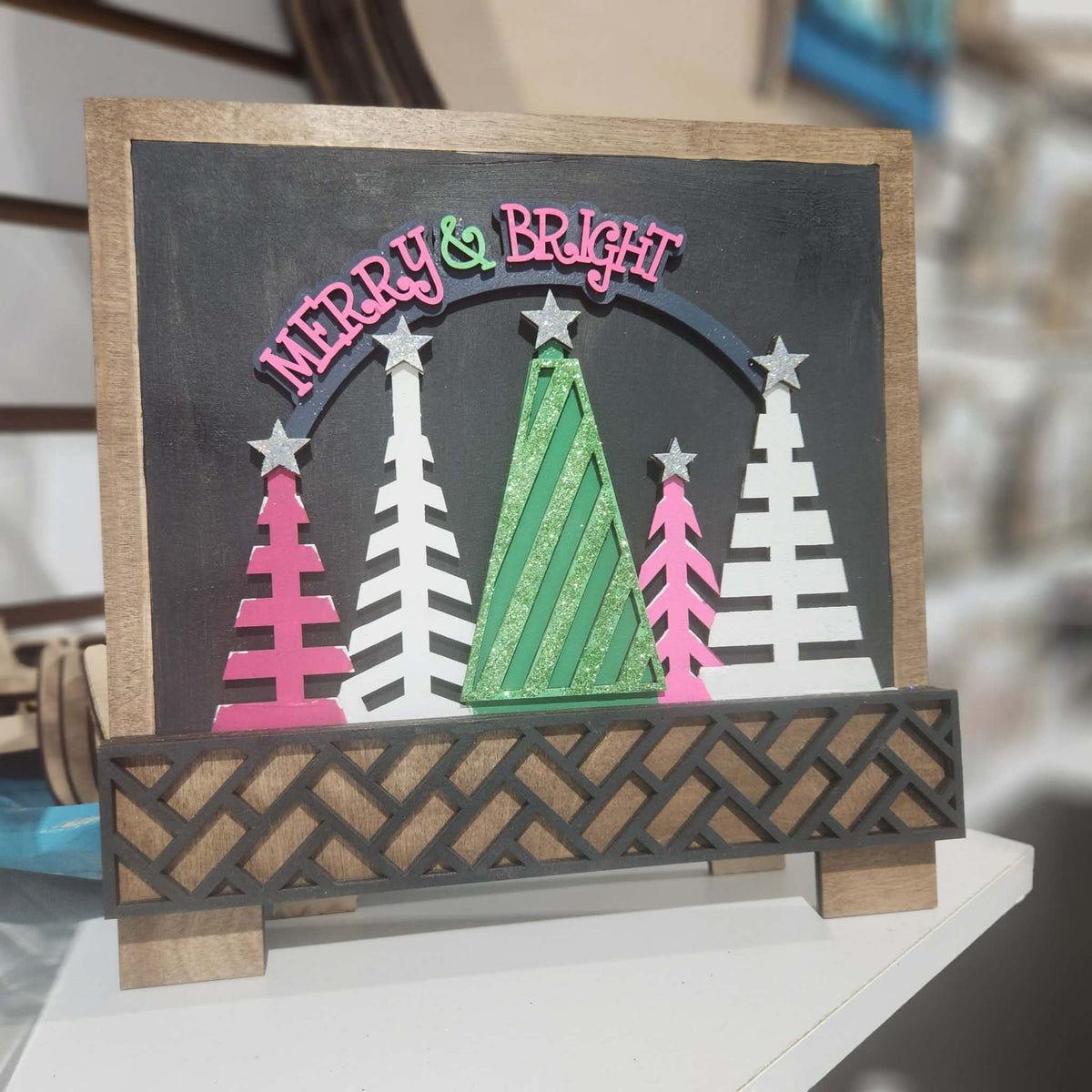 A decorative tabletop holiday sign with a chalkboard-style panel reading “Merry & Bright,” layered paper Christmas trees in pink, white, and green, and a dark woven-pattern front piece on wooden stands.