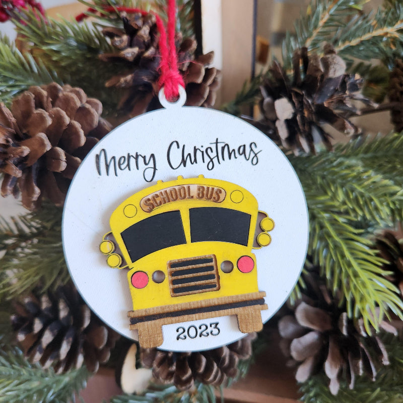 A 3D ornament in the shape of a yellow school bus with 'Merry Christmas' and a date  printed on the side, hanging from a Christmas tree.