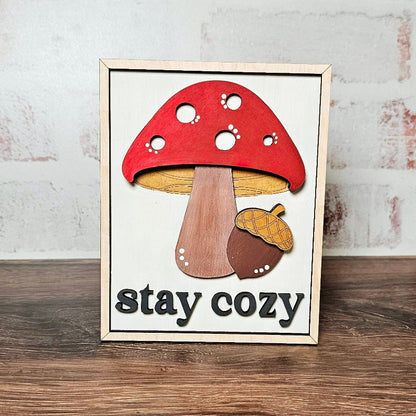 Framed artwork of a red mushroom with 'stay cozy' text on a wooden surface.