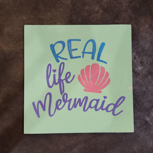 Green sign with 'Real life Mermaid' text and a pink shell on a brown background