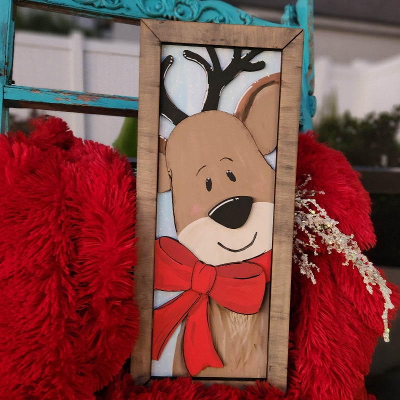 A framed 3D plank featuring a colorful reindeer design with a red bow and white fur, displayed on a red plush surface.