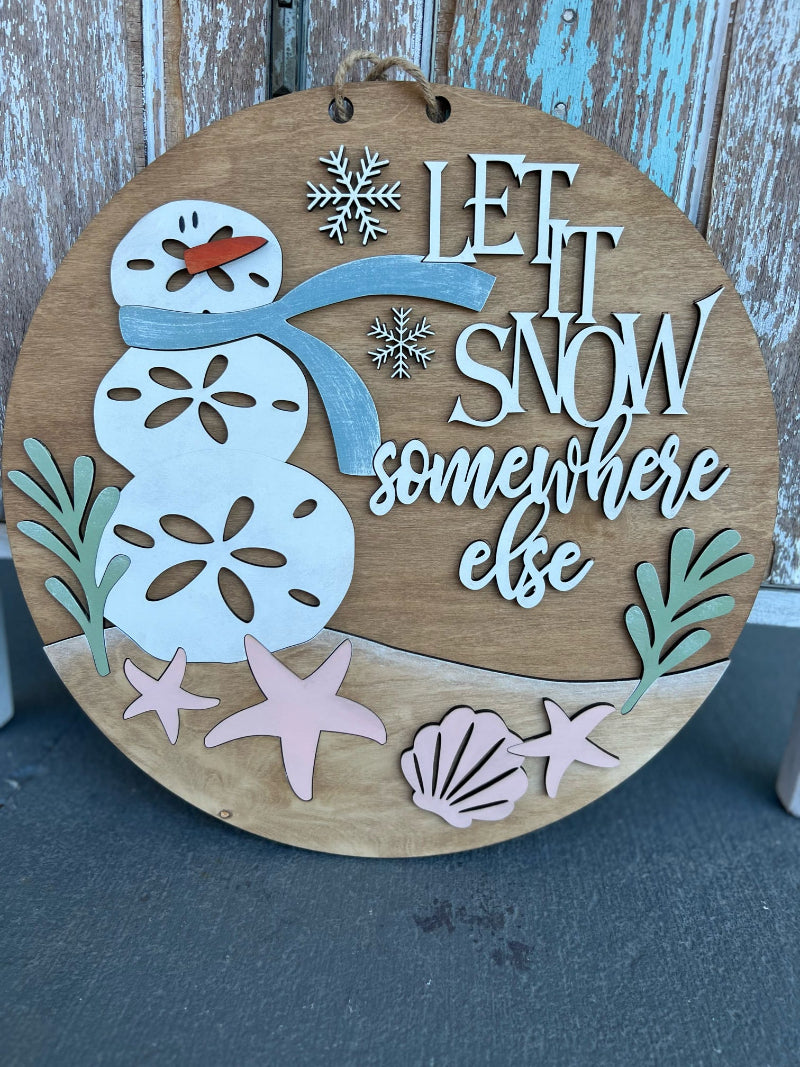 Wooden circular sign with a snowman design and 'Let it snow somewhere else' text, against a wooden background.