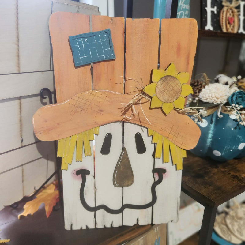 A rustic wooden scarecrow wall decor piece with a white face, yellow hair, a brown hat, and a small sunflower accent, displayed on a shelf in a home decor setting.