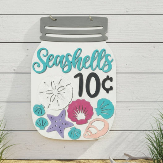 Decorative sign with 'Seashells 10 cents' text and beach-themed graphics on a sandy background.