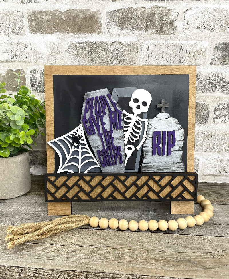 A Halloween-themed wooden sign with a skeleton, tombstones reading 'People Save Me From the Graves' and 'RIP', a spiderweb banner, and a black woven base, displayed on a rustic wood surface with a brick wall and potted plant in the background.