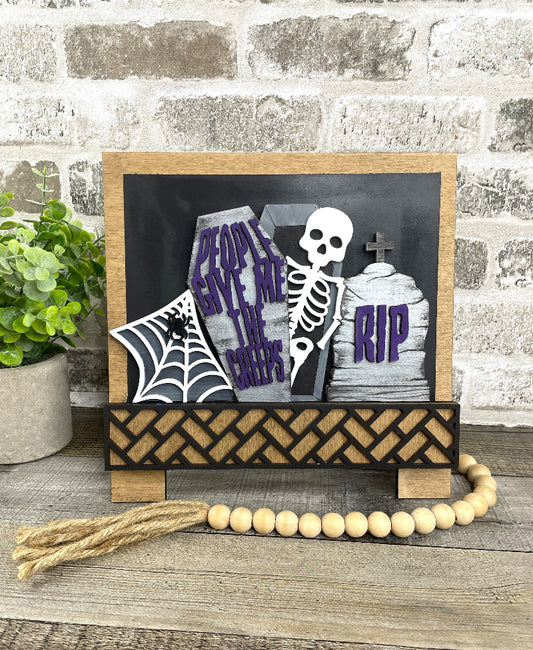 A Halloween-themed wooden sign with a skeleton, tombstones reading 'People Save Me From the Graves' and 'RIP', a spiderweb banner, and a black woven base, displayed on a rustic wood surface with a brick wall and potted plant in the background.