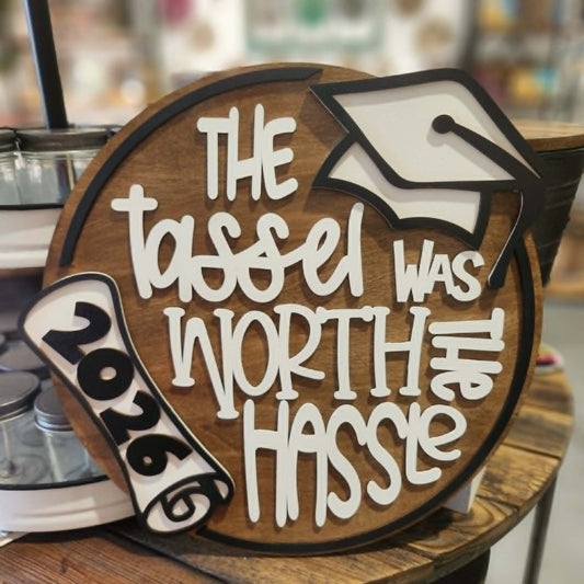wooden background with white letters that says The tassel was worth the hassel. A graduation cap and diploma 