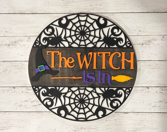 A 3D round door hanger with a witch theme, featuring a witch's hat and a cauldron, set against a wooden background with a spider web design.