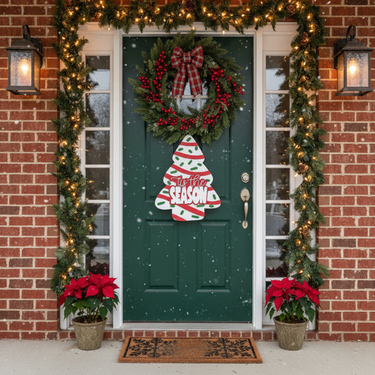 Decorated front door with wreath, tree-shaped sign, and poinsettias on a brick wall.