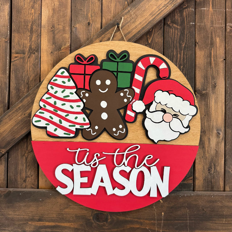 Round wooden sign with Christmas-themed decorations and 'tis the season text on a wooden background