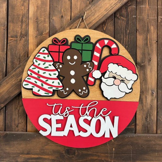 Round wooden sign with Christmas-themed decorations and 'tis the season text on a wooden background