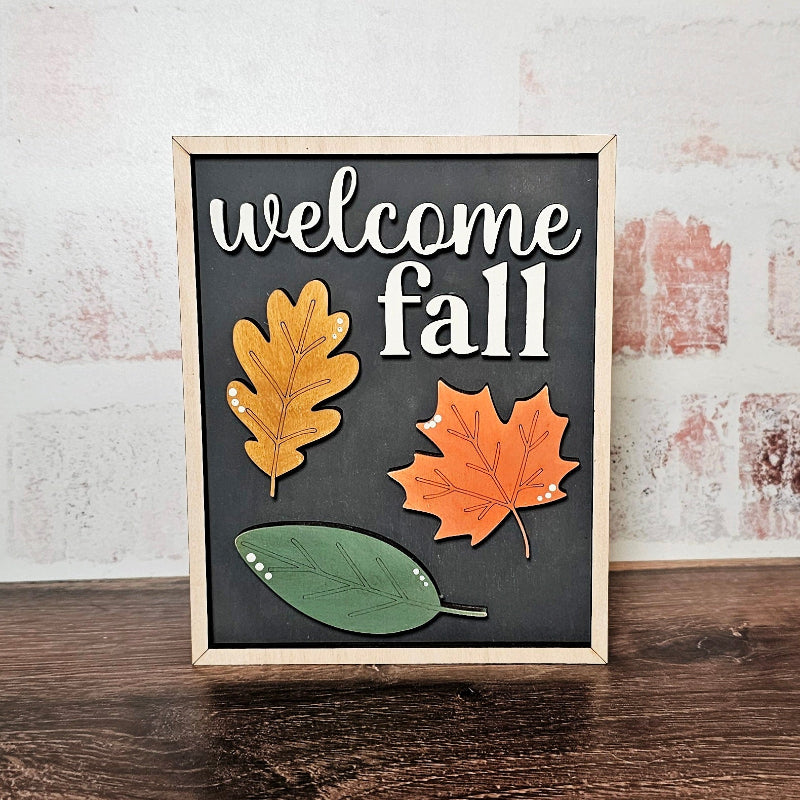 Decorative sign with 'welcome fall' text and autumn leaves on a wooden surface.