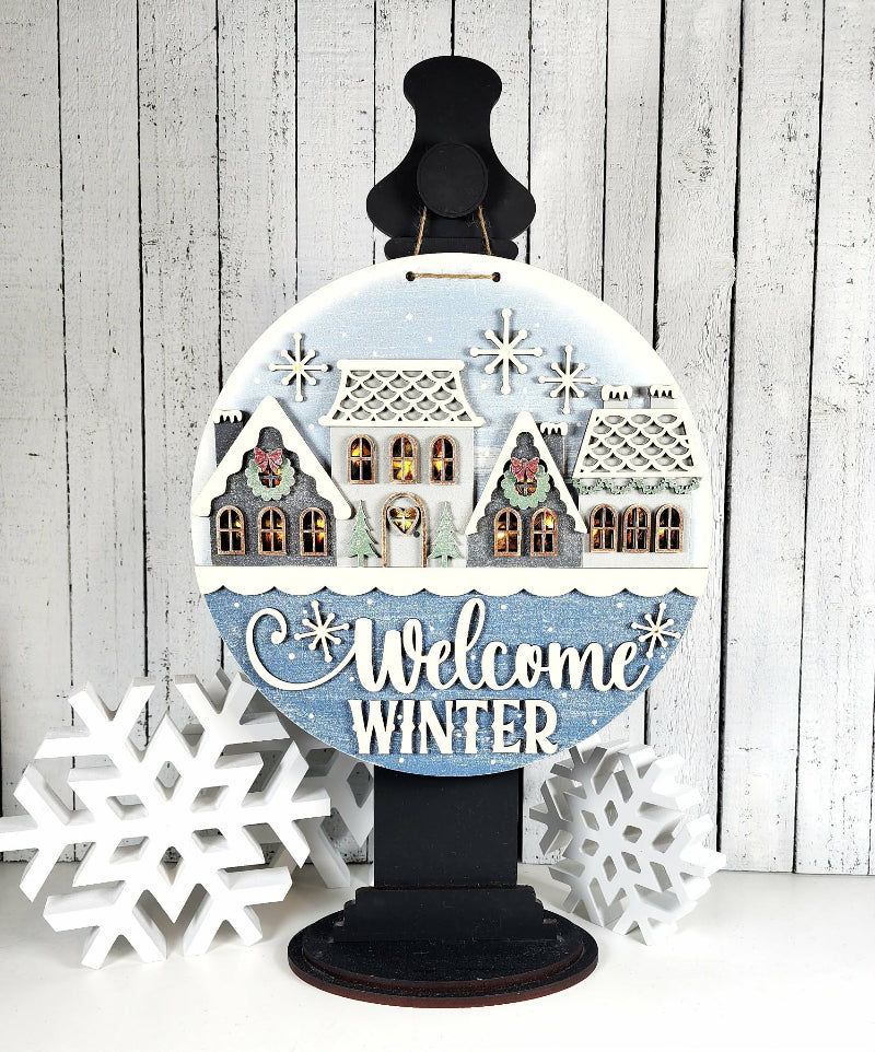 Decorative sign with winter scene and 'Welcome Winter' text on a wooden background