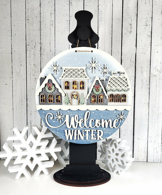 Decorative sign with winter scene and 'Welcome Winter' text on a wooden background