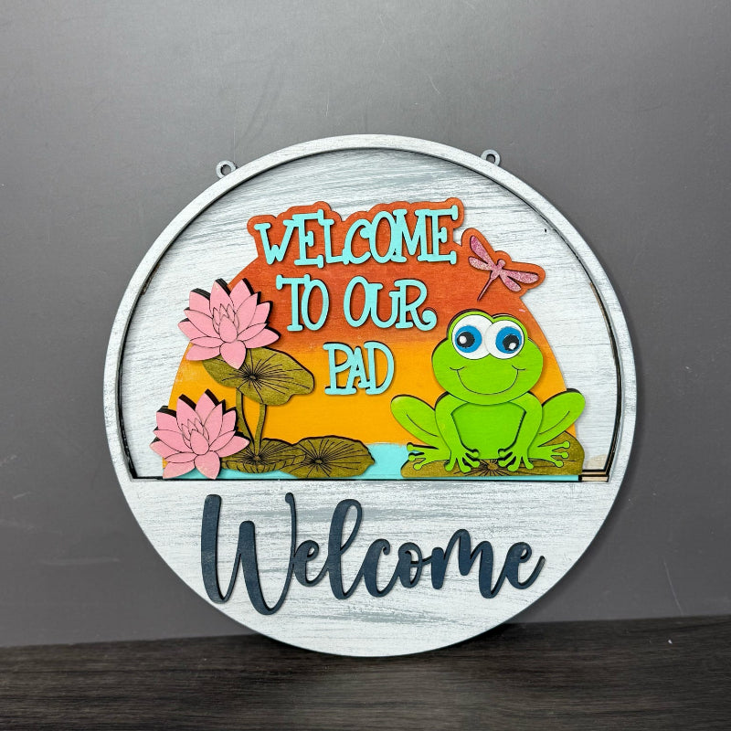 Round decorative sign with a frog and floral design, featuring the text 'Welcome to our pad' on a gray background.