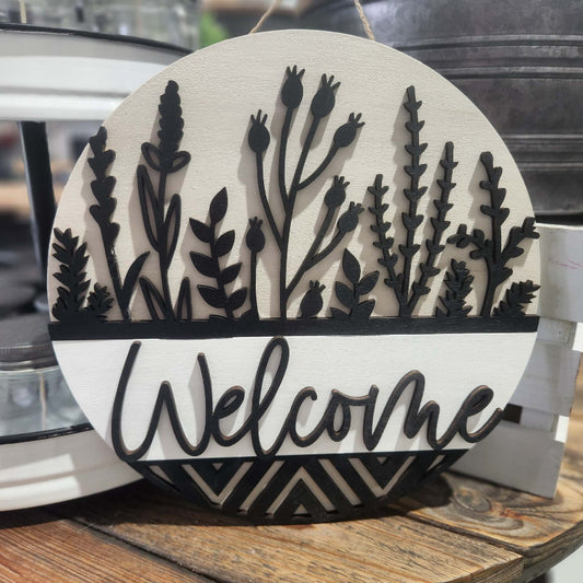 Decorative sign with black flower silhouettes and 'Welcome' text on a wooden surface.