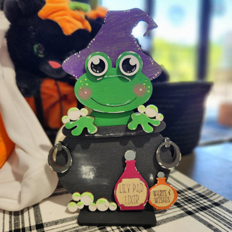 Halloween-themed decoration with a green frog in a witch hat on a black cauldron.