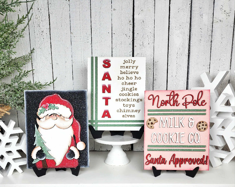 Three Christmas-themed signs on a wooden surface with decorative elements.