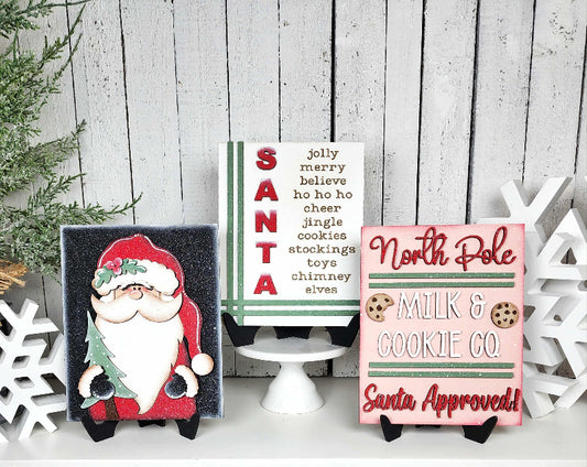 Three Christmas-themed signs on a wooden surface with decorative elements.