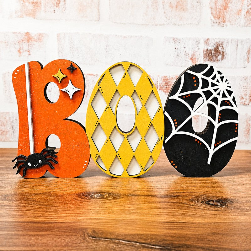Three Halloween-themed wooden letters spelling BOO, displayed on a wooden tabletop against a white brick wall. The letter B is orange with a black spider and white stars; the O is yellow with a diamond lattice pattern; the R is black with a white spiderweb and small orange dots.