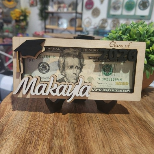 Wooden frame with graduation cap and 'Class of 2026' text, featuring a dollar bill design with 'Makayla' on a wooden surface.