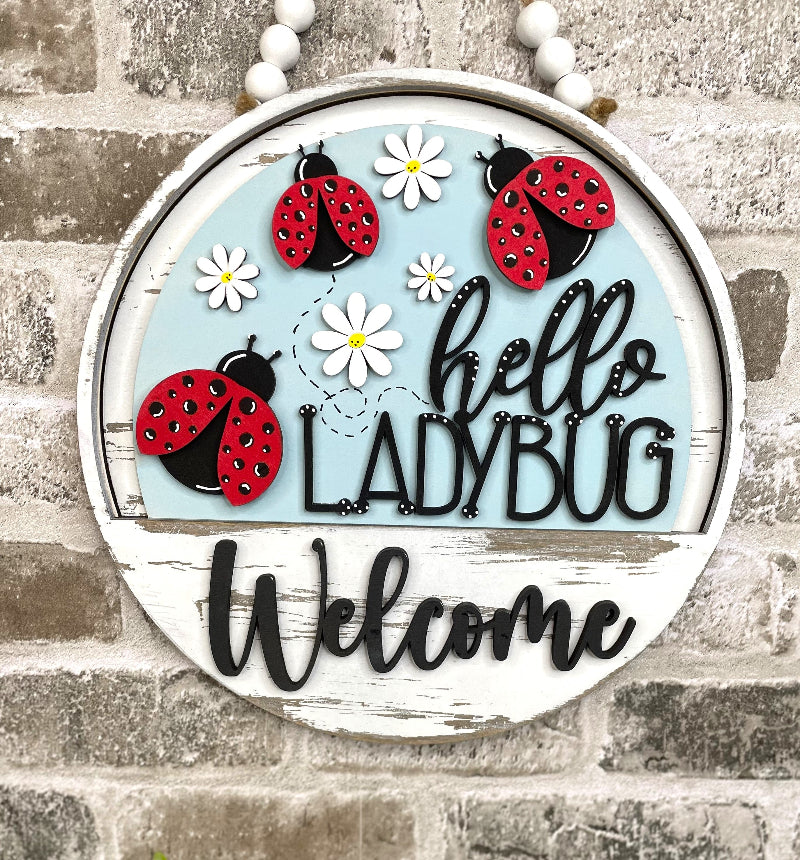 Hello Ladybug interchangeable wooden Welcome sign insert with three red ladybugs, white daisies and black lettering on a light blue background.