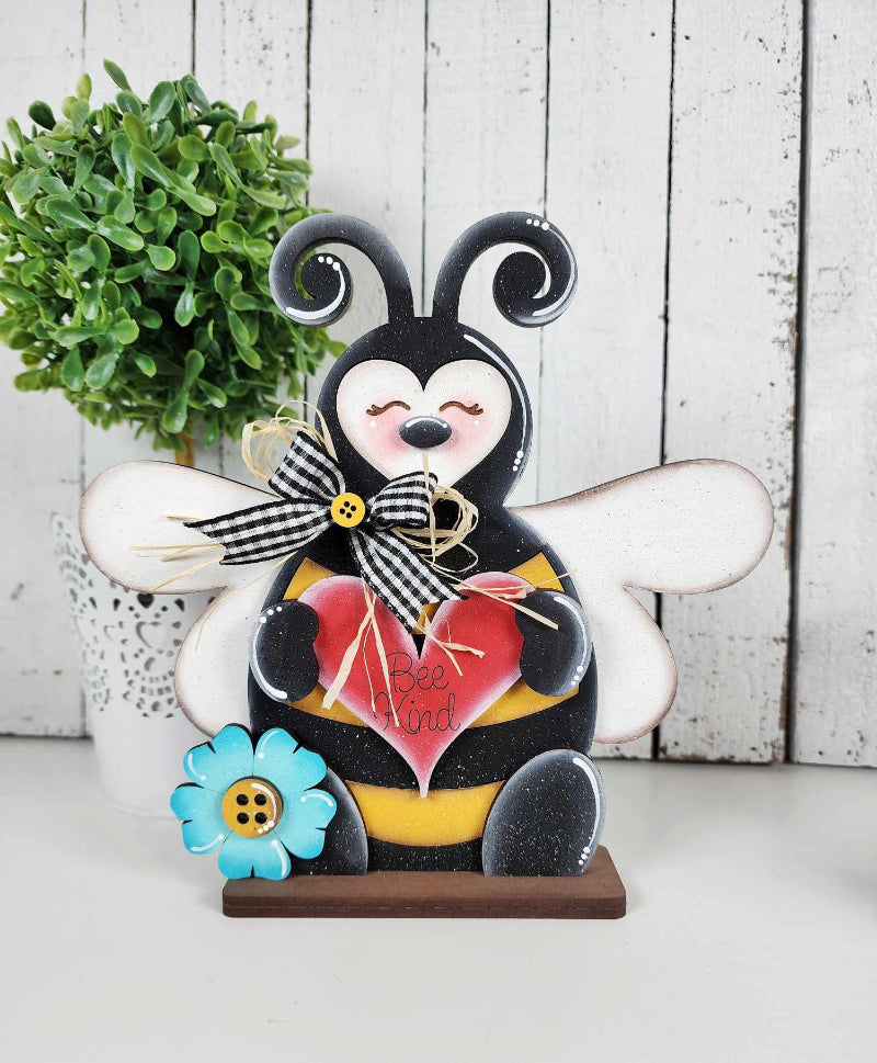A decorative honeybee figurine with a heart, bow, and flower design, sitting on a shelf.