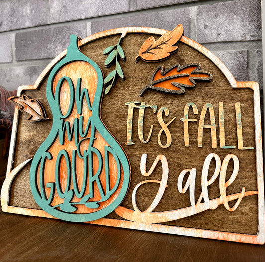 A wooden 3D rectangle sign with the text 'Oh My Gourd It's Fall Y'all' cut out in various fonts and colors, along with decorative leaf and gourd shapes.