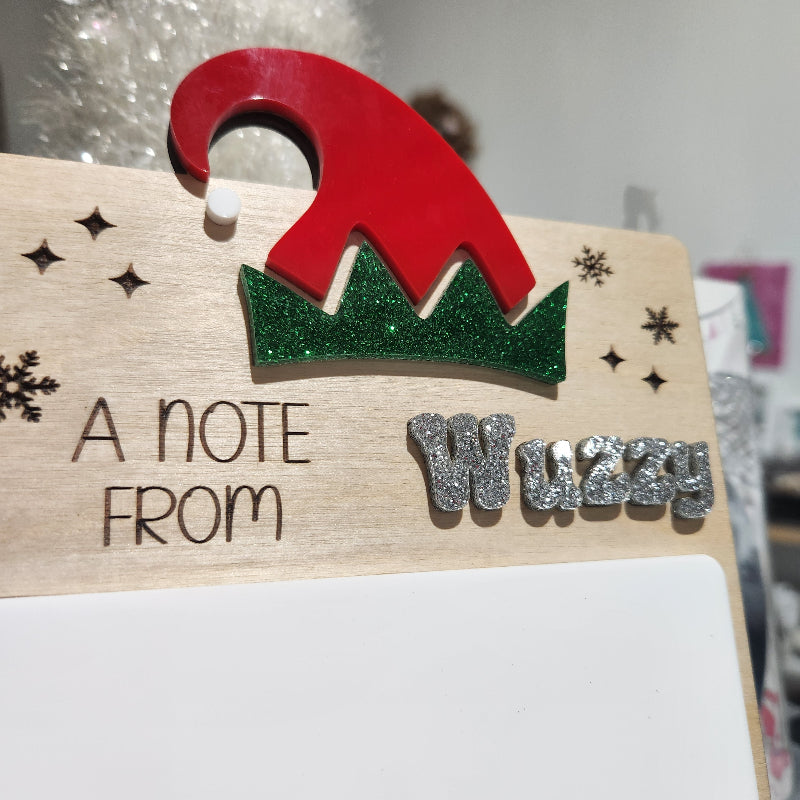 Wooden message board with red and green tree design and 'A Note from' text on a blurred background