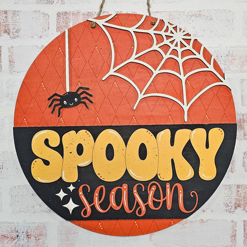 Round Halloween wall sign with orange and black sections, white spiderweb cutout, black spider, and the words “SPOOKY season” in yellow and orange lettering, hanging against a white brick wall.