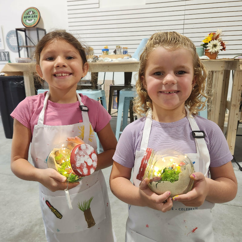 Two children wearing aprons holding decorated eggs in a craft room.