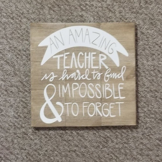 A square wooden artwork with the text 'An Amazing Teacher is hard to Find' in a whitewashed black font, placed on a floor.
