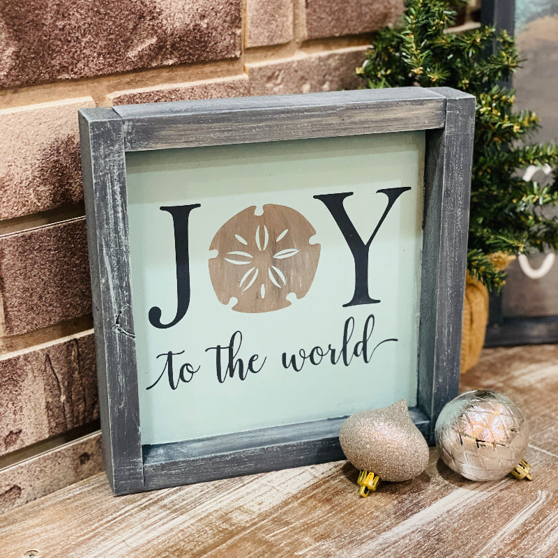 Decorative sign with 'Joy to the world' text and sand dollar design, placed on a wooden surface with Christmas decorations.