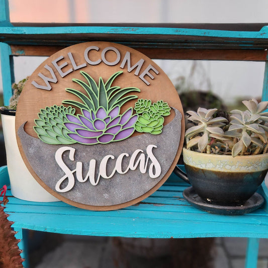 A round wooden sign with the words Welcome Succas around the edge and 3D succulents in the middle
